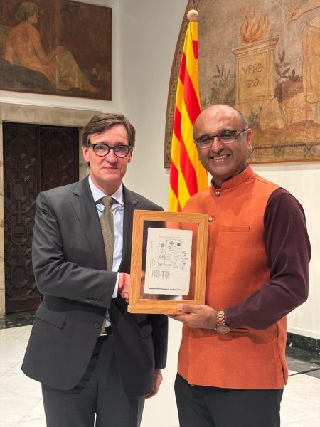 Two men standing and shaking hands and holding a certificate. The one on the left is wearing glasses and a suit and the one on the right is also wearing glasses and wearing a sleeveless orange Nehru jacket.