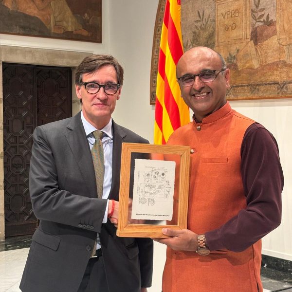 Two men standing and shaking hands and holding a certificate. The one on the left is wearing glasses and a suit and the one on the right is also wearing glasses and wearing a sleeveless orange Nehru jacket.